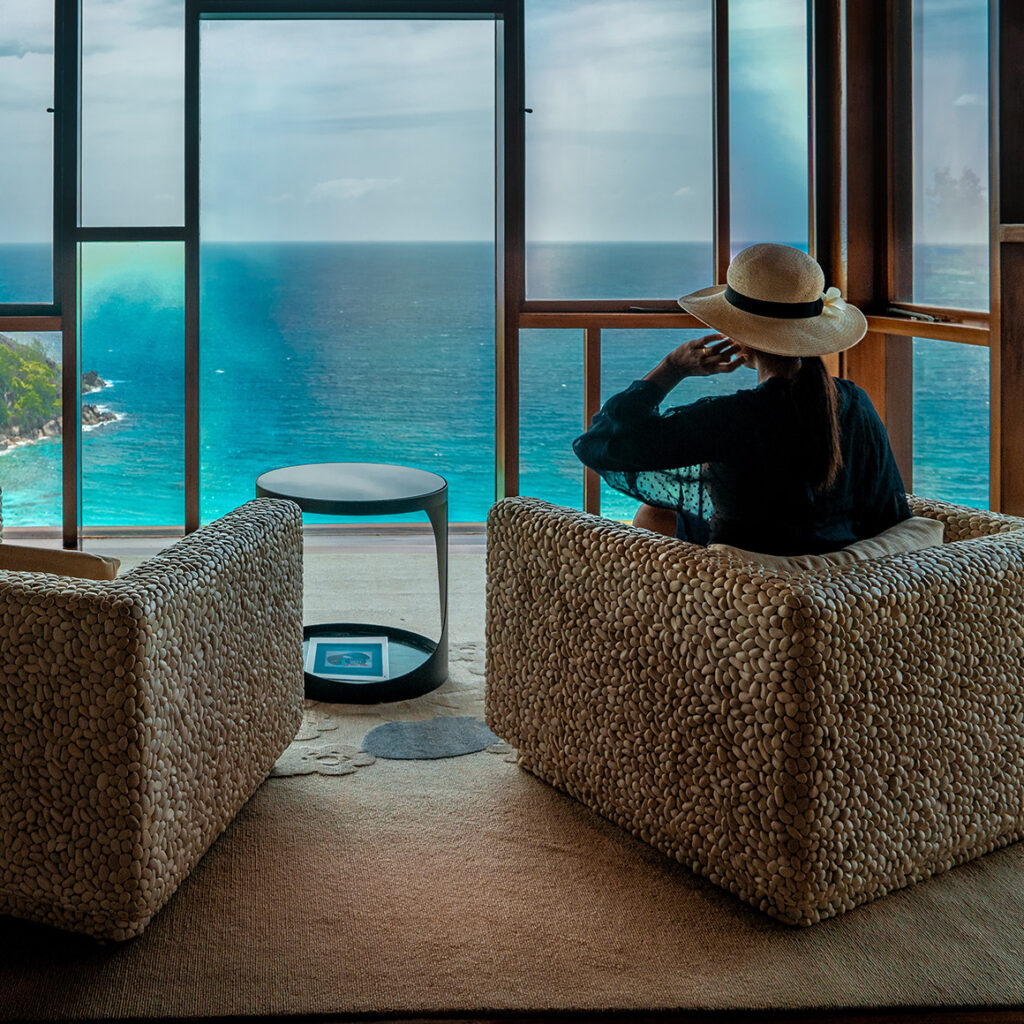 a woman sitting on a chair looking pensively out the window in front of her which overlooks a vast ocean